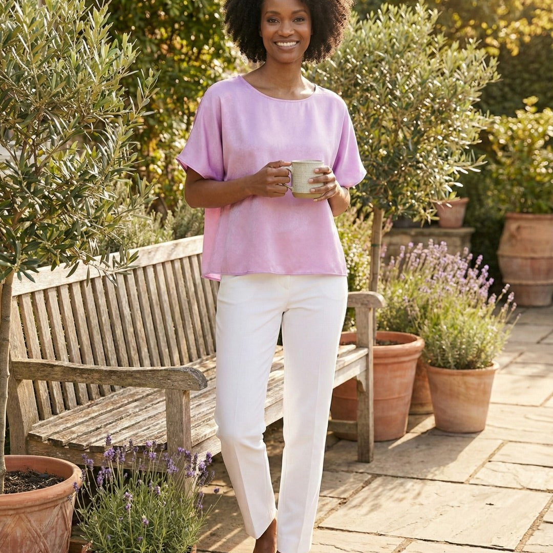 Woman in a pink shirt and white pants standing in a garden holding a mug.