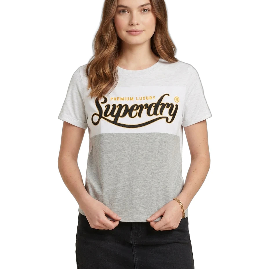 A woman modeling a Superdry colorblock t-shirt with a white and heather gray top half, and a dark gray bottom half. The front features a large brown 'Superdry' logo with 'PREMIUM LUXURY' written above it.