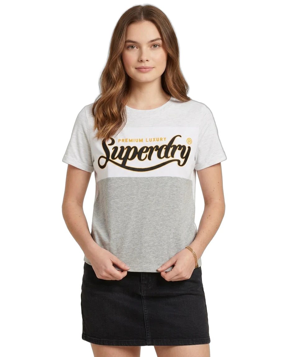 A woman modeling a Superdry colorblock t-shirt with a white and heather gray top half, and a dark gray bottom half. The front features a large brown 'Superdry' logo with 'PREMIUM LUXURY' written above it.