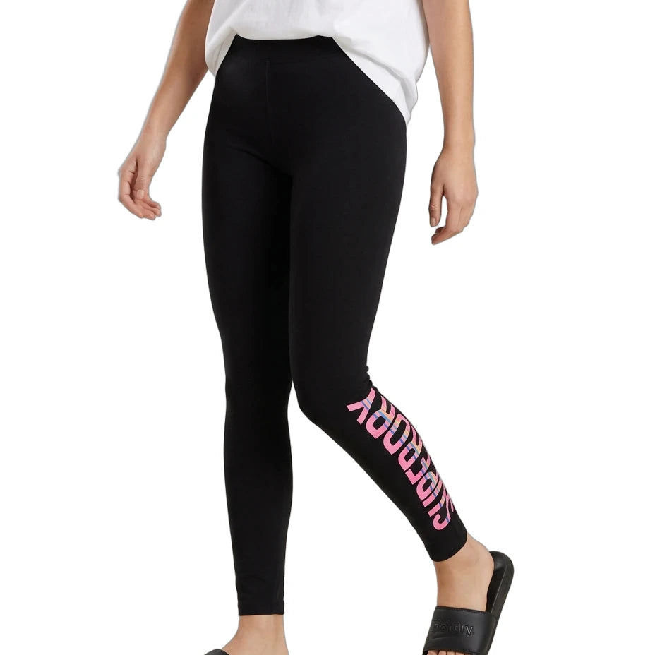 A model wearing black leggings with a pink 'SUPERDRY' logo printed vertically down the left leg, styled with a white t-shirt and black slide sandals against a plain white background.