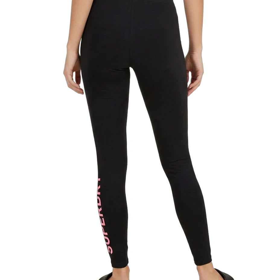 A model shown from the back wearing black leggings with a pink 'SUPERFRONT' logo printed vertically down the left leg, paired with black slide sandals on a white background.