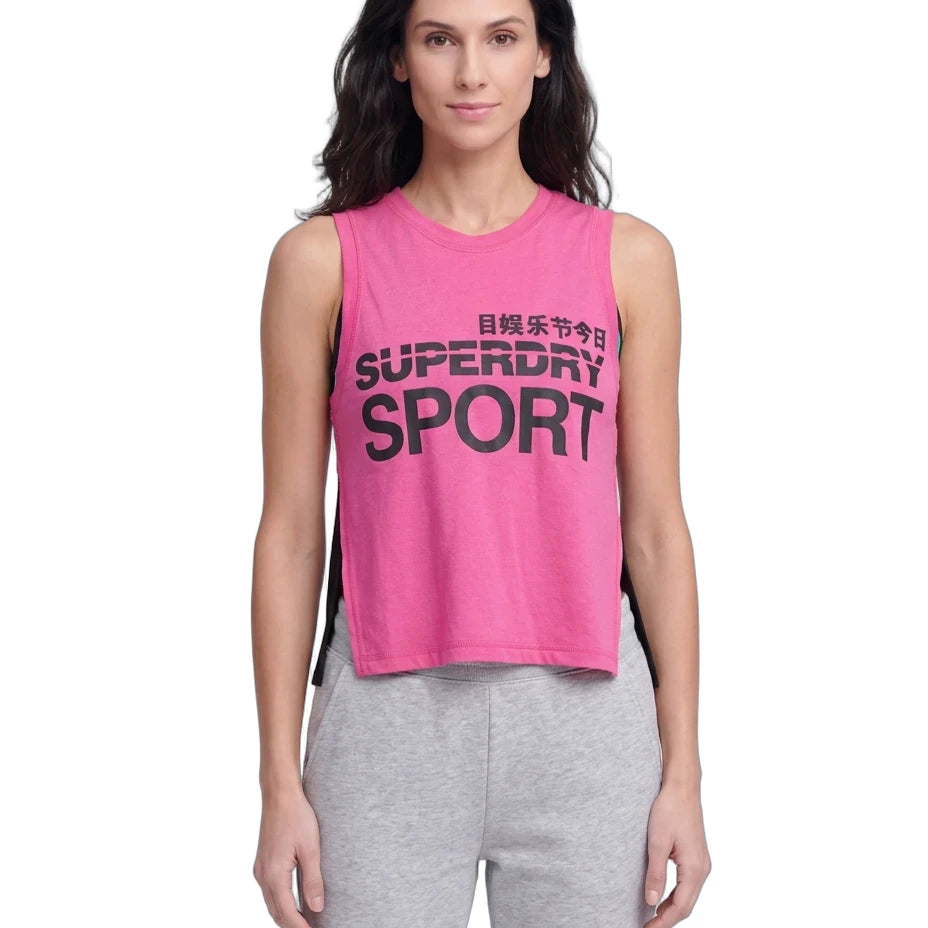 A woman modeling a bright pink sleeveless crop top with black 'SUPERDRY SPORT' text on the front, paired with light gray sweatpants, against a plain white background.