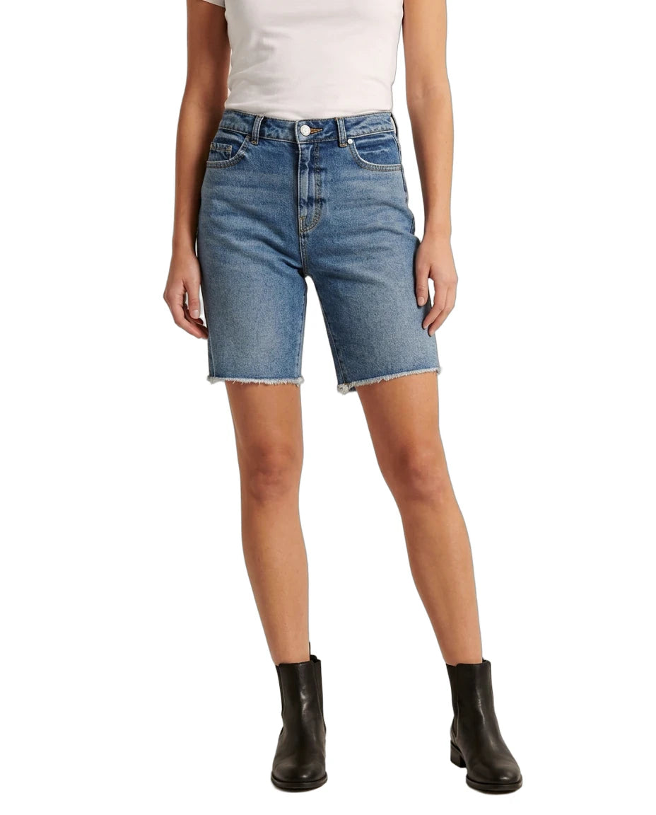A model wearing medium-wash blue denim shorts with a raw, frayed hem, paired with a white top and black ankle boots, shown against a plain white background.