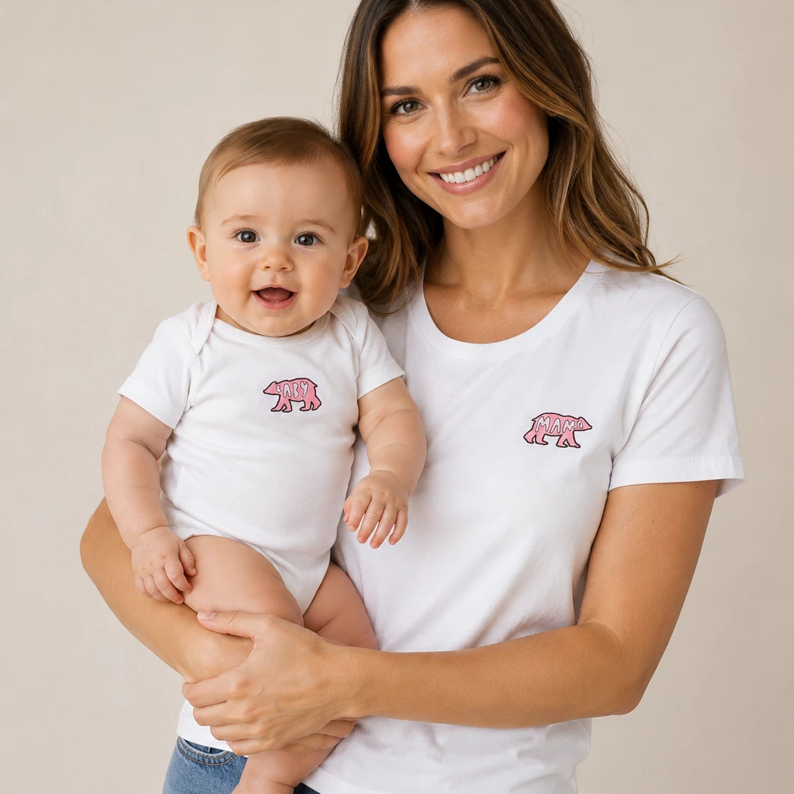 A smiling woman holding a baby, both wearing matching white short-sleeve bodysuits with a small pink polar bear graphic on the chest. The woman is also wearing blue jeans, and the background is a clean, neutral beige studio backdrop.