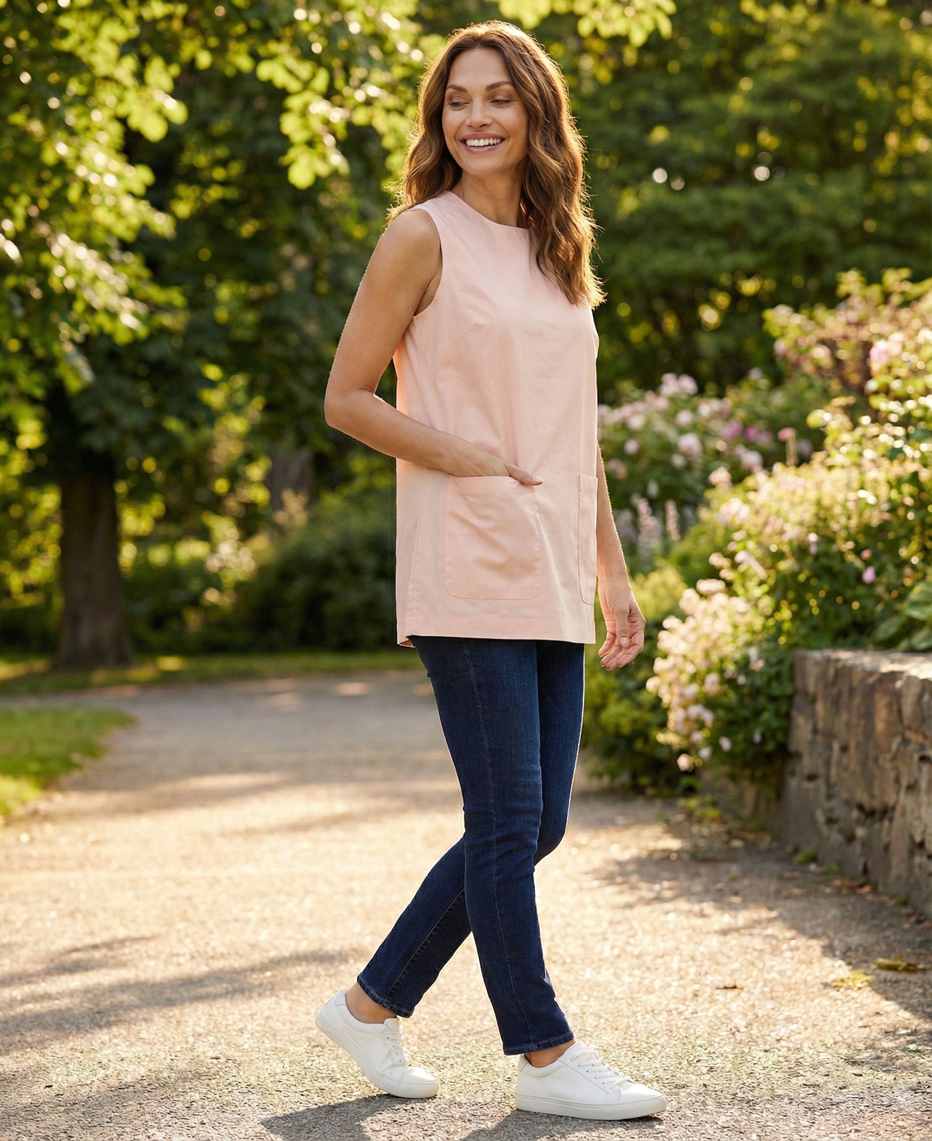 Woman walking outdoors in a park wearing a light pink sleeveless top and dark blue jeans.
