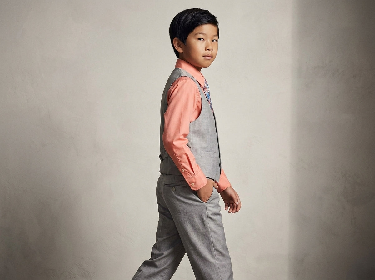 A young boy wearing a gray vest and matching gray trousers over a coral pink long-sleeve shirt, standing against a textured gray wall.