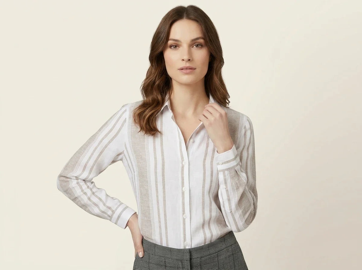 Brunette woman modeling a white and beige vertical-striped button-up linen blouse tucked into grey plaid trousers against a neutral background.