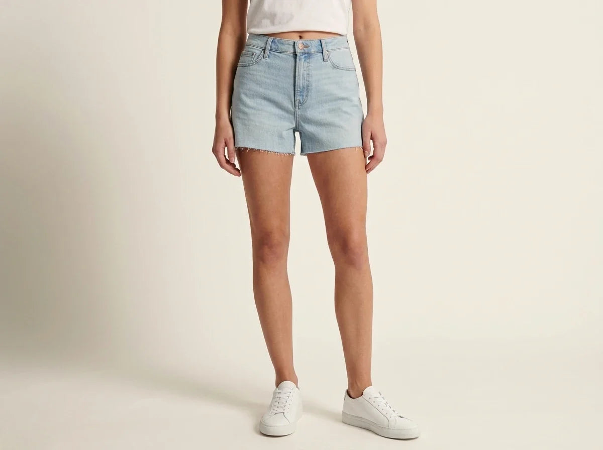 Cropped front view of a woman wearing light-wash high-rise denim shorts with raw hems, a white t-shirt, and white low-top sneakers.