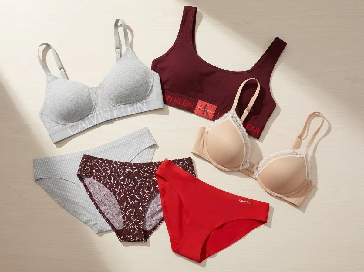 Flat-lay collection of Calvin Klein women's bras, bralettes, and panties in grey, burgundy, beige lace, and red on a neutral surface.