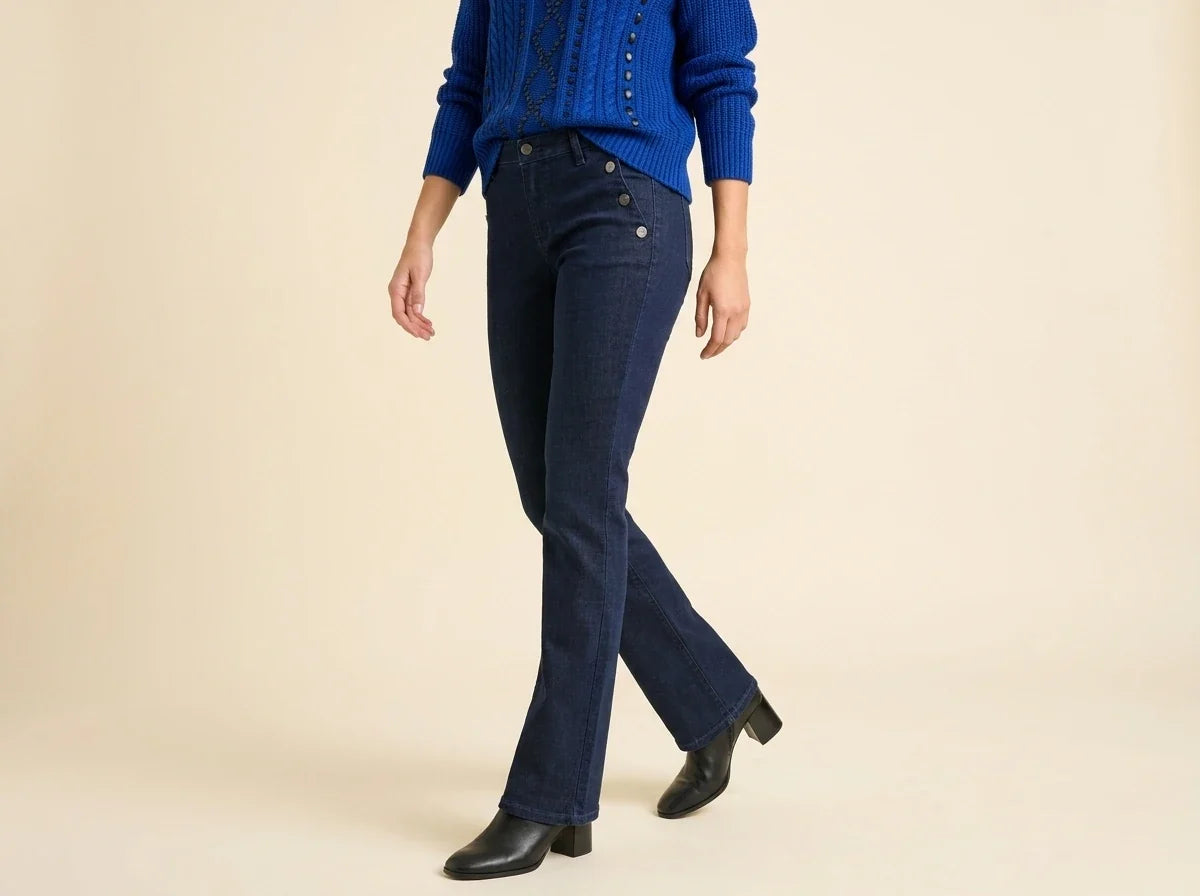 A model wearing dark blue flared jeans, a blue knit sweater, and black ankle boots, shown from the chest down against a solid beige studio background.