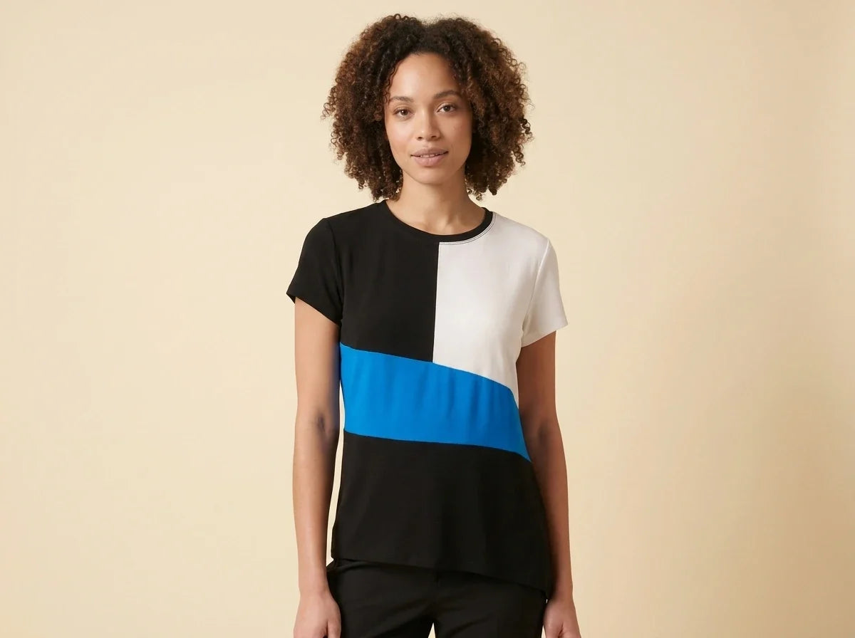 A woman with curly brown hair wearing a short-sleeve colorblock top with black, white, and bright blue blocks, paired with black pants, standing against a solid beige background.