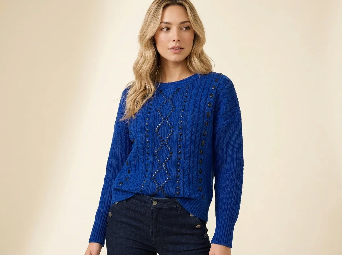A woman modeling a royal blue cable-knit sweater with decorative dark beadwork down the center front and dark buttons along the sleeves, paired with dark jeans against a warm beige studio background.