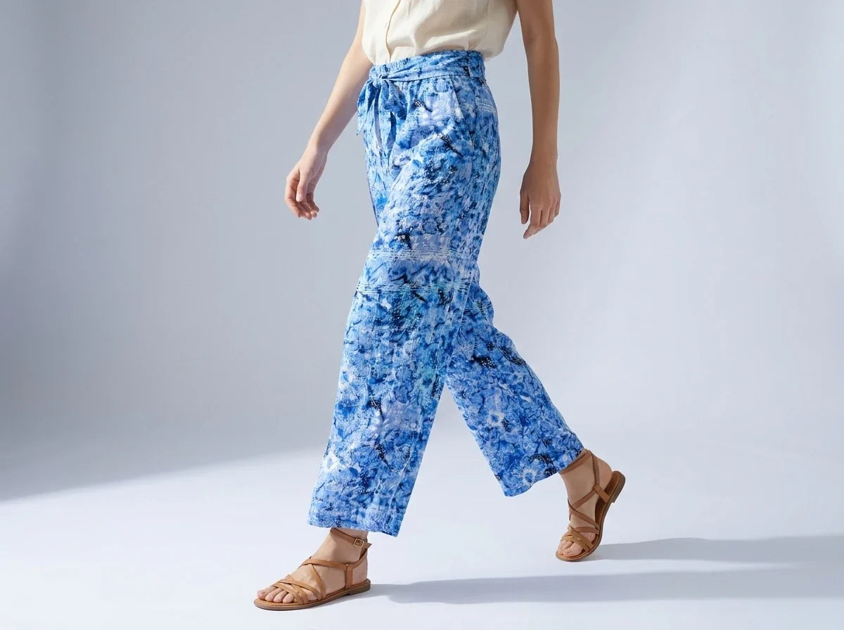 A model wearing blue and white abstract patterned wide-leg pants with a drawstring waist, styled with a cream top and brown leather sandals against a light gray studio background.