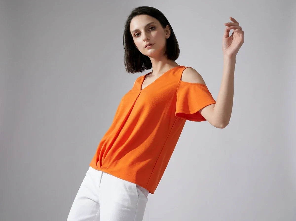 A woman modeling a bright orange cold-shoulder top with white pants, posed against a neutral gray studio background.