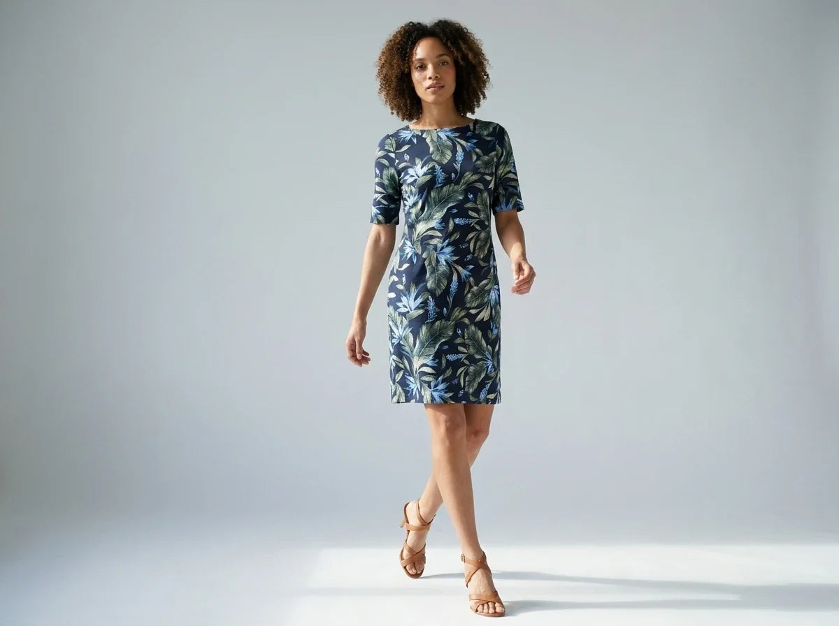 A woman modeling a knee-length short-sleeve dress with a blue and green tropical leaf pattern, paired with tan strappy high-heeled sandals, against a light gray studio background.