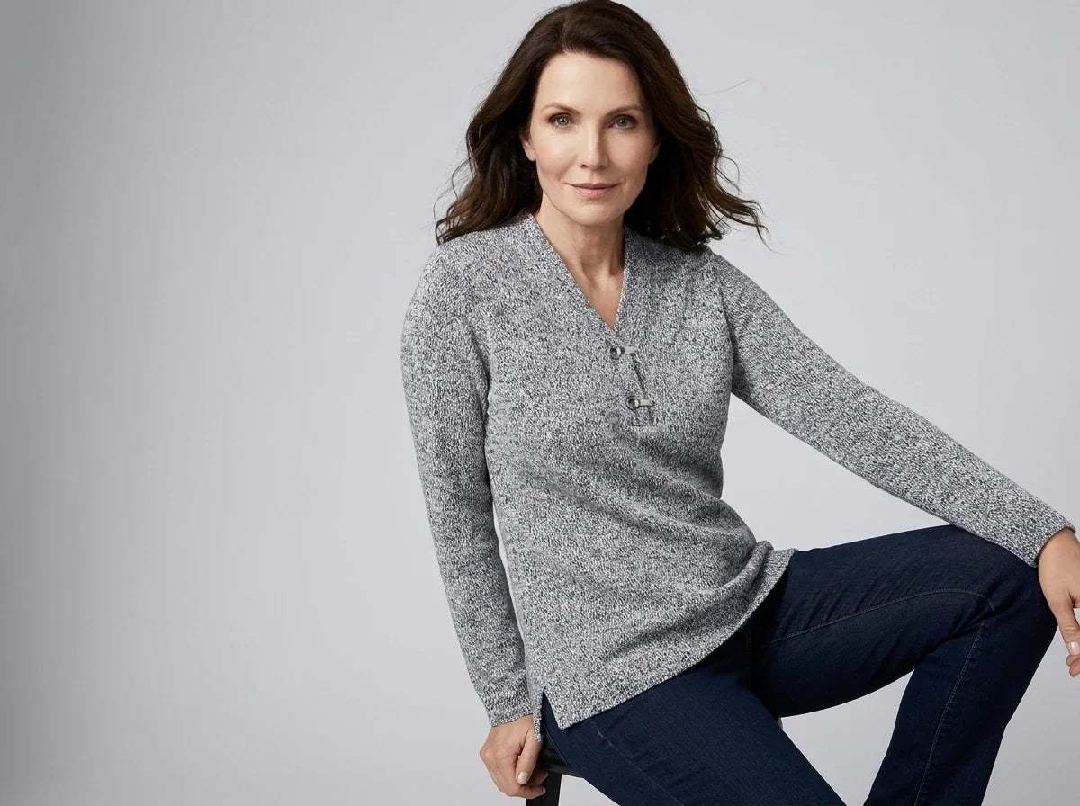 A woman wearing a gray marled long-sleeve henley sweater and dark blue jeans, posing against a light gray studio background.