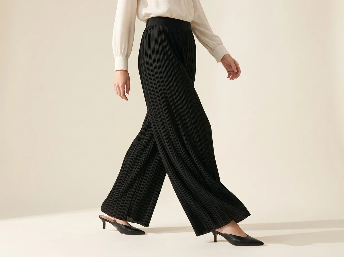 Cropped view of a woman walking in black pleated wide-leg palazzo pants paired with black pointed-toe kitten heel mules and a cream blouse.