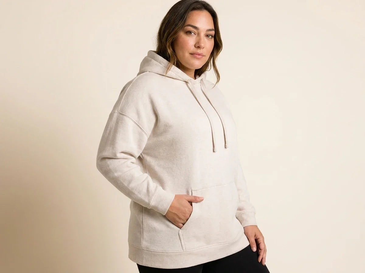 Brunette woman modeling a plus-size oatmeal heather fleece hoodie sweater with a front kangaroo pocket and black leggings.