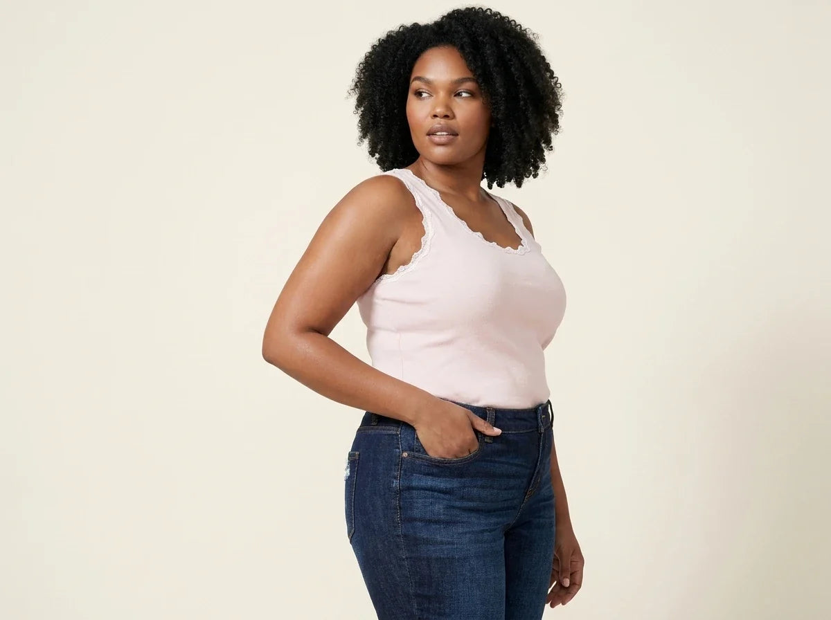 Curvy woman with curly black hair modeling a light pink lace-trimmed scoop-neck tank top tucked into dark wash jeans.