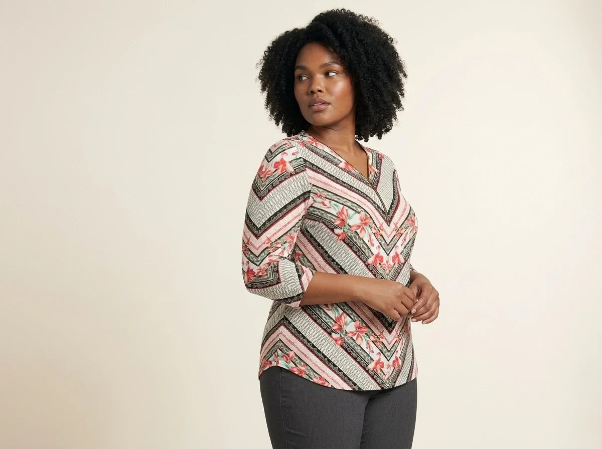 Curvy woman with curly black hair modeling a plus-size chevron and floral print three-quarter sleeve tunic top paired with grey trousers.