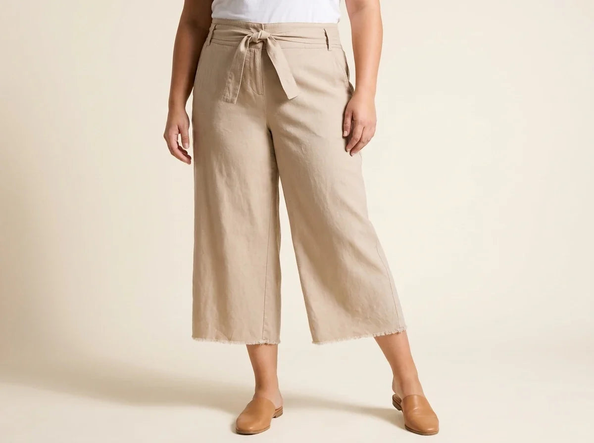 Cropped view of a woman wearing high-waisted beige linen tie-front wide-leg culottes with raw fringe hems and tan leather mules.