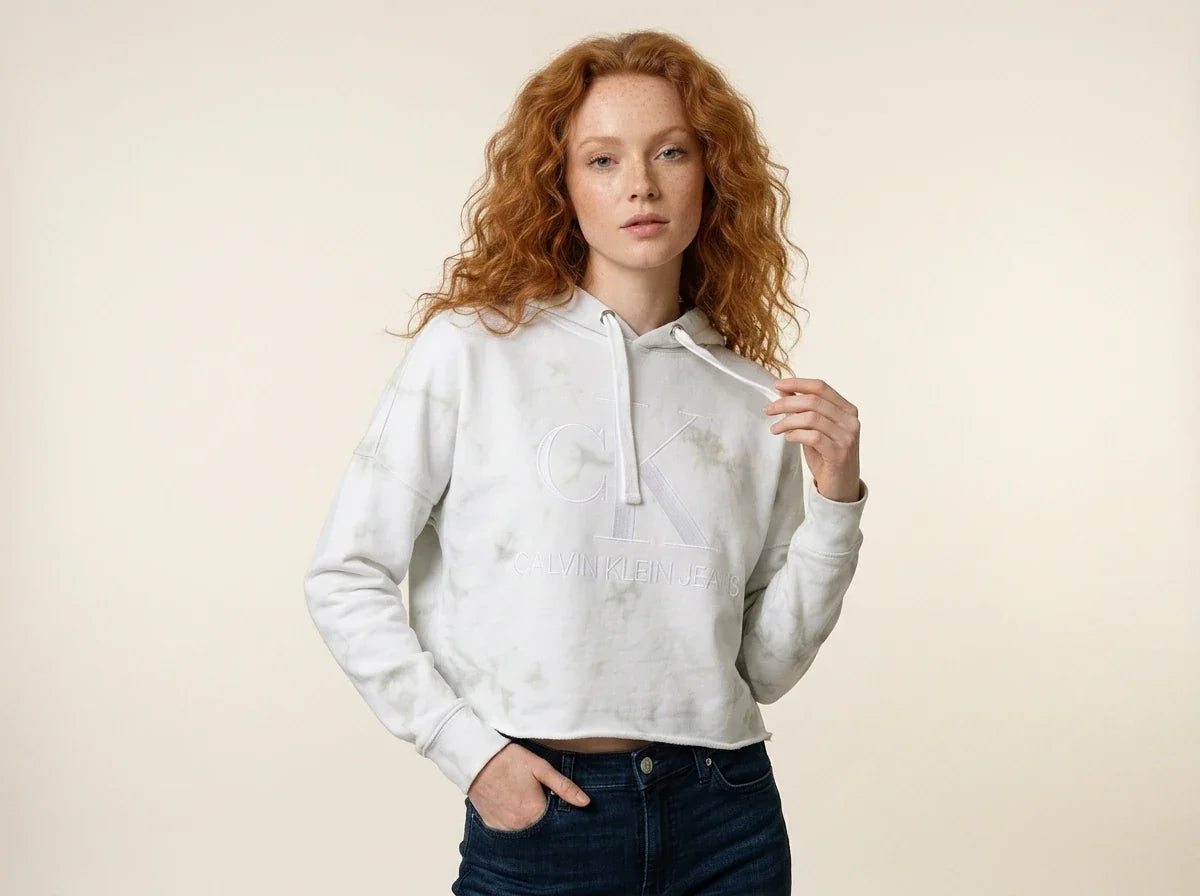 Woman with curly red hair modeling a Calvin Klein Jeans light green tie-dye cropped hoodie paired with dark wash jeans.