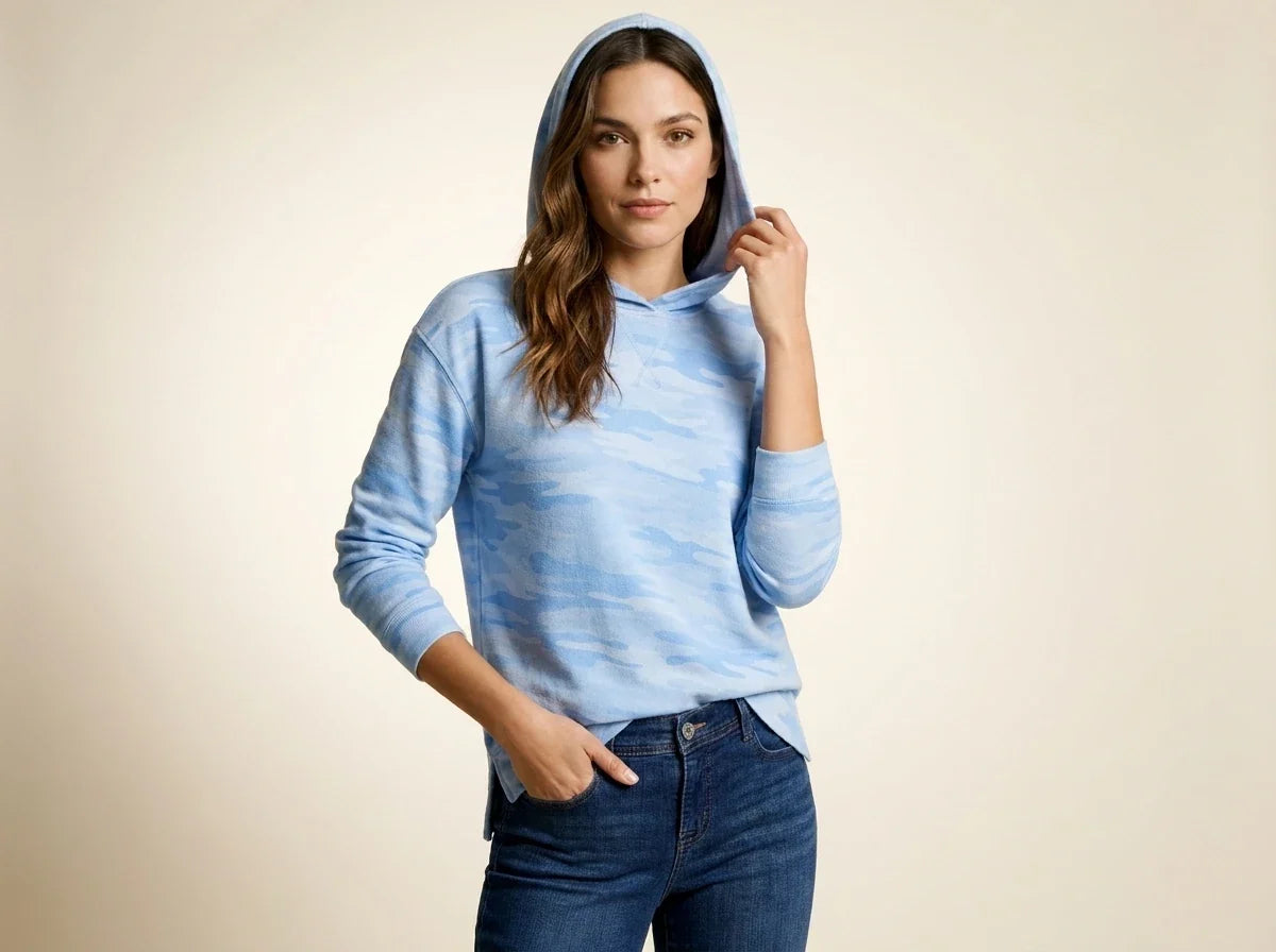Brunette woman modeling a light blue camo print hooded pullover sweatshirt with side slits paired with dark wash jeans.