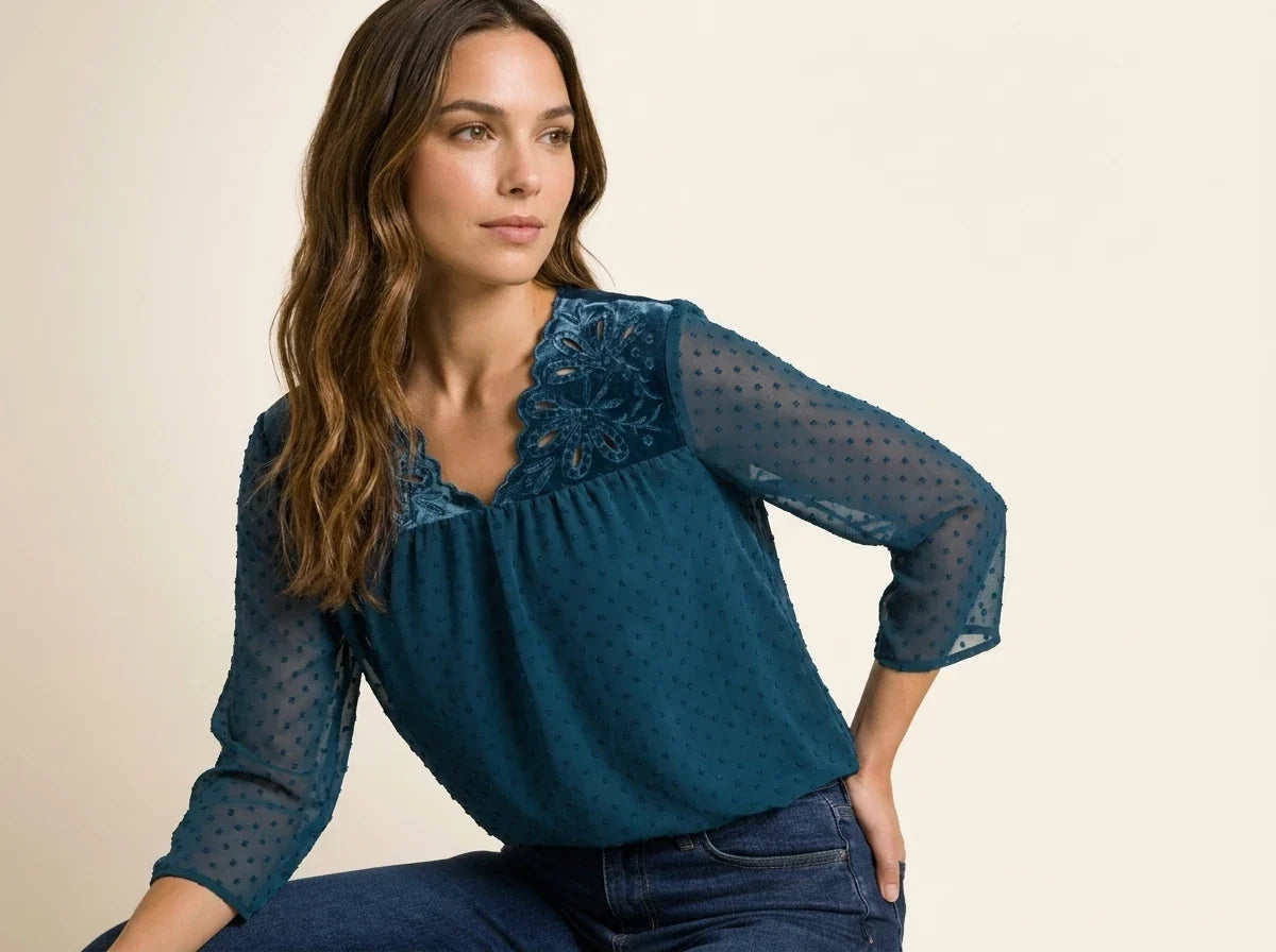 Brunette woman modeling a teal blue Swiss dot chiffon blouse featuring a velvet floral cutout neckline paired with dark wash jeans.