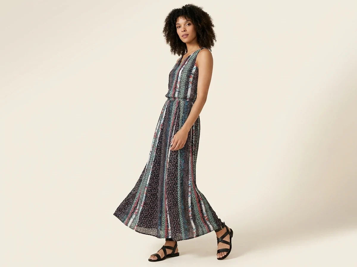 Woman with curly brown hair modeling a sleeveless floral and vertical stripe maxi dress paired with black strappy flat sandals.