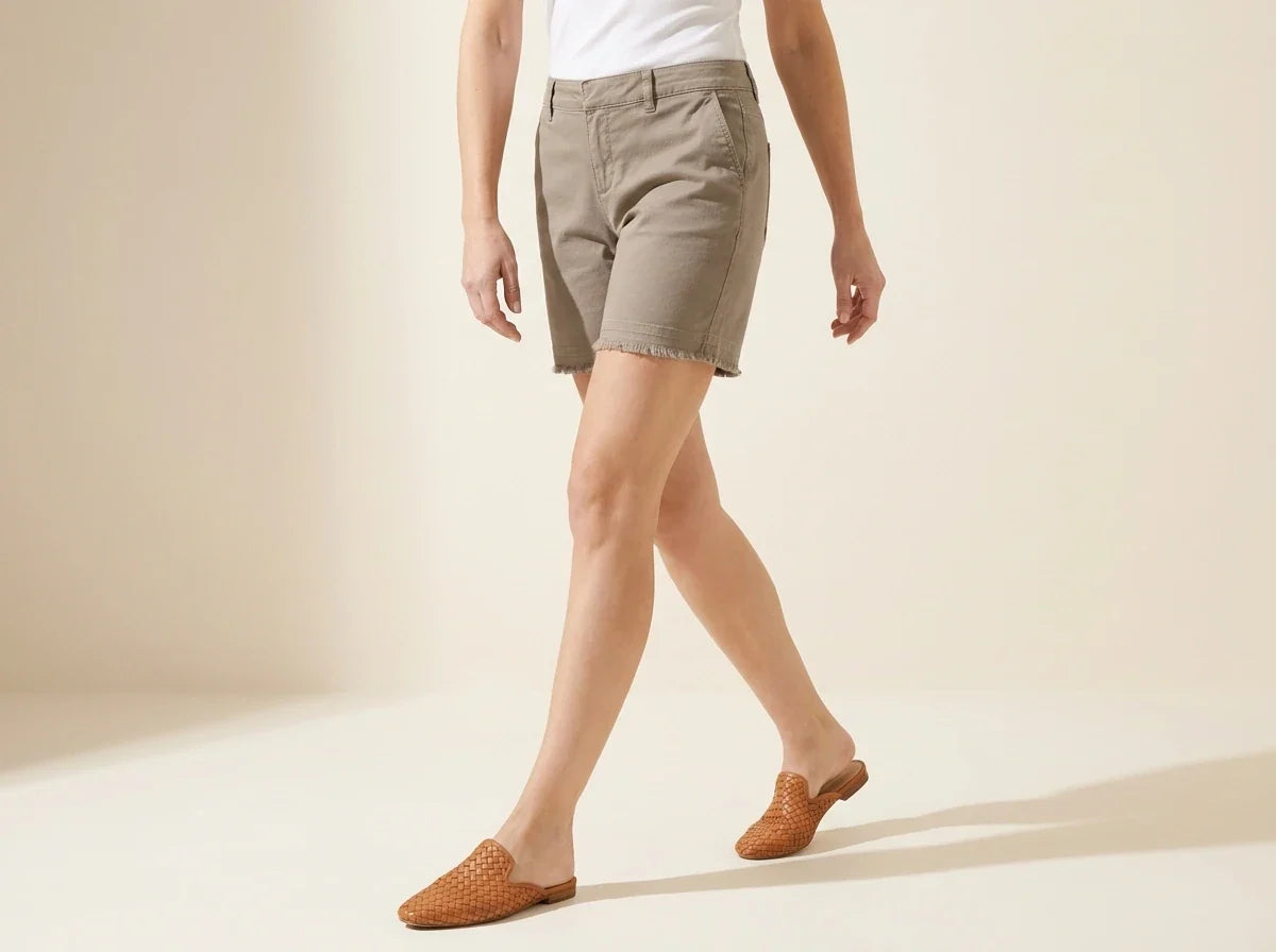 Cropped view of a woman wearing petite taupe khaki stretch shorts with raw fray hem and cognac woven leather mule flats.