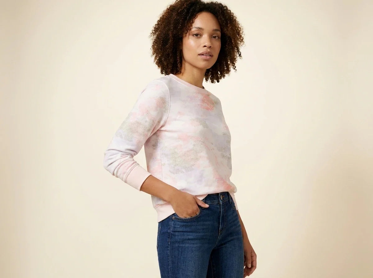 Woman with curly brown hair modeling a pastel tie-dye crewneck fleece sweatshirt paired with medium-wash blue jeans.