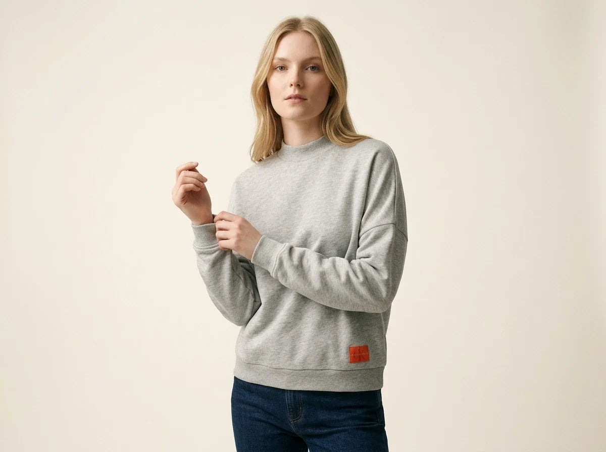 Blonde woman modeling a Calvin Klein heather grey drop-shoulder sweatshirt with an orange logo patch, paired with dark wash jeans.