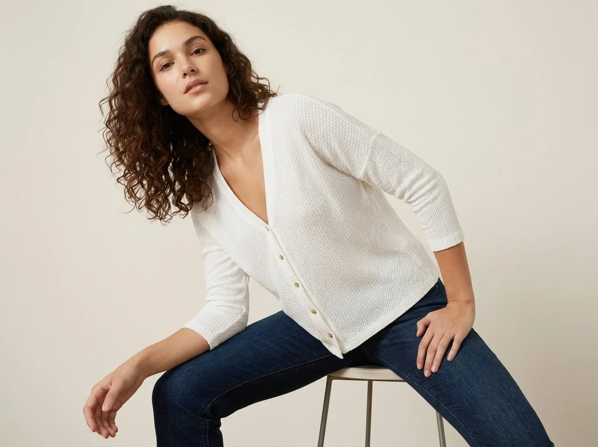 A woman with curly brown hair wearing a white textured knit cardigan and dark blue jeans, sitting on a metal stool against a plain beige studio background.