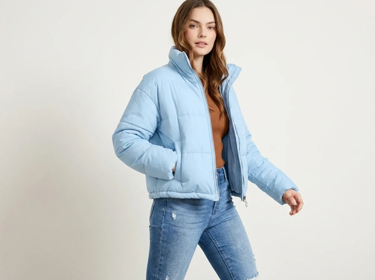 A woman modeling a light blue puffer jacket, styled with a brown top and blue jeans, against a plain off-white studio background.