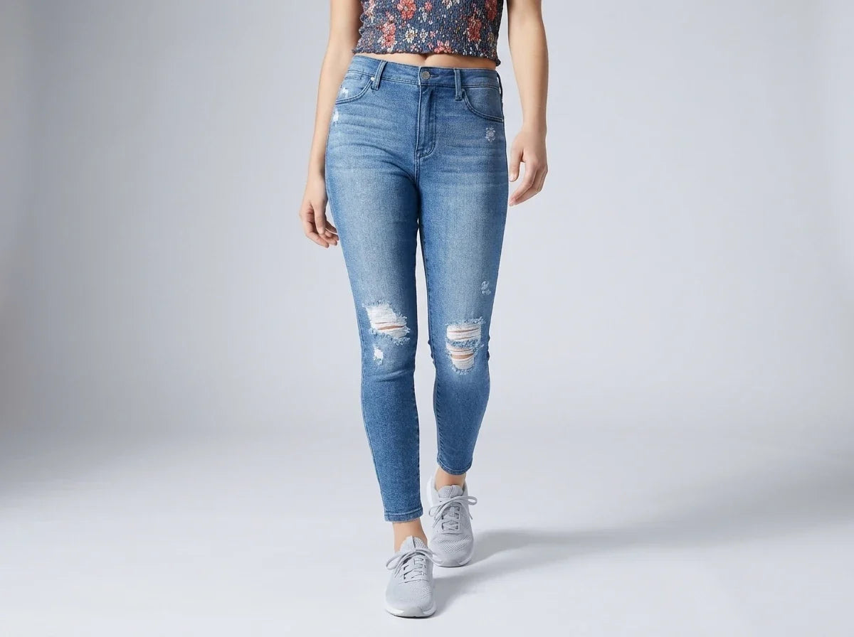 A model wearing light blue distressed skinny jeans with ripped details on the thighs and knees, paired with a patterned crop top and gray sneakers, shown against a light gray studio background.