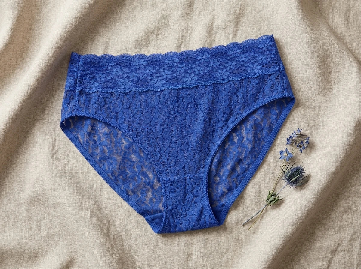Blue lace high-waisted briefs laid flat on a beige linen fabric background, styled with small dried blue flowers in the corner.