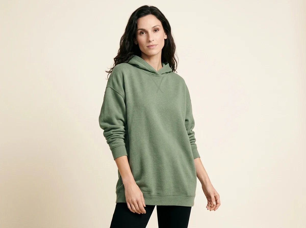 Brunette woman modeling an oversized sage green fleece hoodie sweater with cross-stitch collar detailing and black leggings.