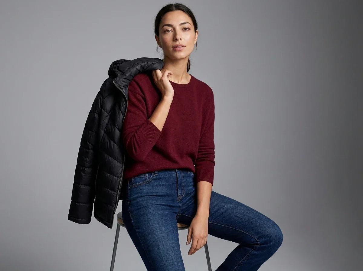 A woman seated on a chair against a gray studio background, wearing a burgundy knit sweater and blue jeans with a black puffer jacket draped over her shoulder.