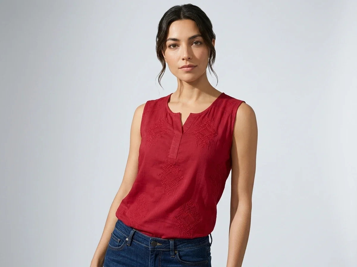 A woman modeling a sleeveless red top with a split neckline and tonal embroidered details, paired with blue jeans, against a light gray studio background.