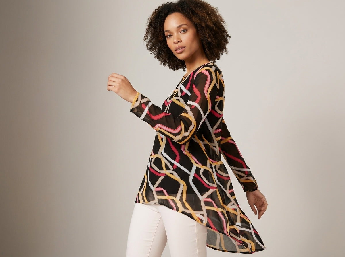 A woman modeling a long-sleeve black tunic top with a colorful abstract chain-link pattern in yellow, pink, and white, paired with white pants against a neutral studio background.