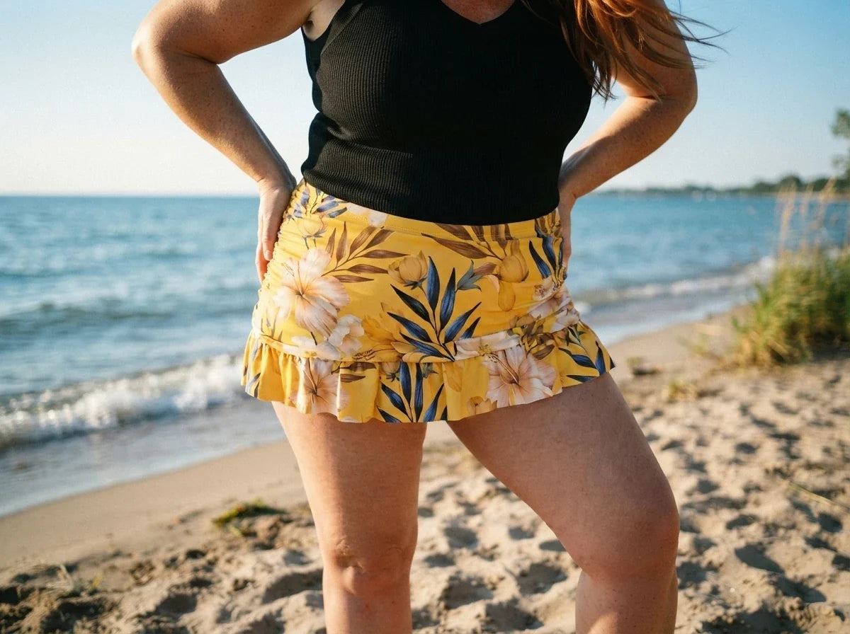 A woman standing on a sandy beach wearing a black ribbed tank top and a yellow ruffled mini skirt with a tropical floral print, with the ocean in the background.