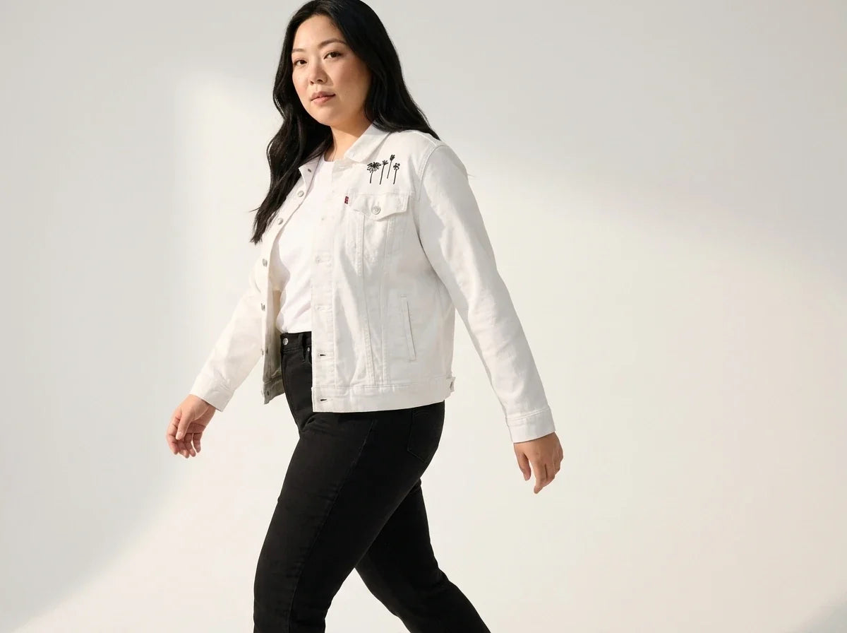 A woman modeling a white denim jacket with black embroidered details on the chest, paired with black pants, against a plain white studio background.