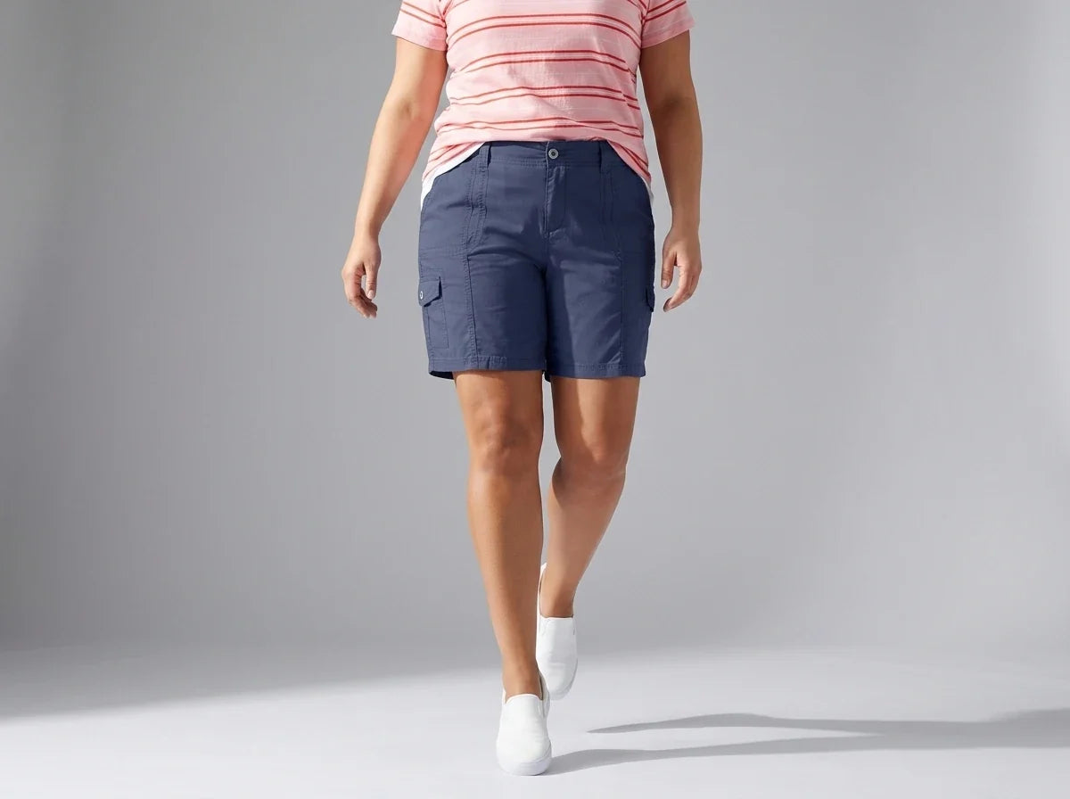 A model wearing navy blue cargo shorts, a pink and white striped t-shirt, and white socks walking against a light gray studio background.