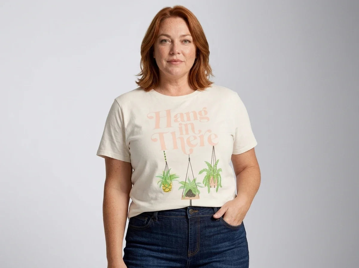 A woman with red hair wearing a cream-colored graphic t-shirt with the words 'Hang in There' and illustrations of hanging plants, paired with dark blue jeans, against a light gray studio background.