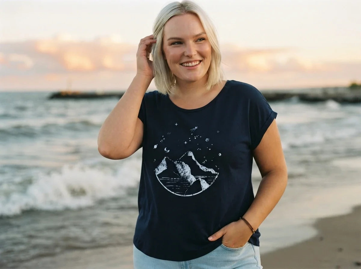 A smiling blonde woman wearing a navy blue graphic t-shirt with a white moon and mountain design, standing on a beach during sunset.