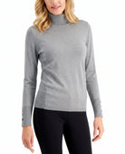 A woman wearing a light gray long-sleeve mock neck sweater with decorative buttons on the cuffs, paired with black pants, against a plain white background.