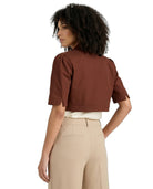 A woman shown from the back wearing a cropped brown short-sleeve top with rolled cuffs, paired with beige trousers and a white undershirt, against a plain white background.