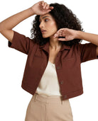 A woman wearing a cropped brown short-sleeve button-front jacket over a cream top and beige trousers, posed against a plain white studio background.