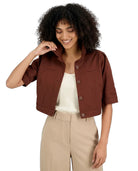 A woman with curly black hair wearing a brown cropped short-sleeve button-up jacket over a cream top and beige trousers, photographed against a plain white background.