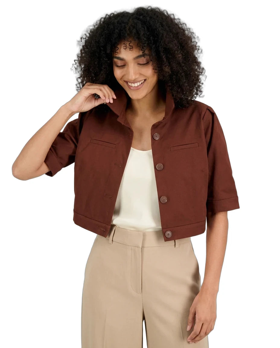 A woman with curly black hair wearing a brown cropped short-sleeve button-up jacket over a cream top and beige trousers, photographed against a plain white background.